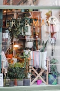 A display window of a store named Bert & Mays showcases an assortment of plants, decorative pillows, and other home decor items. Several potted plants are visible, arranged on shelves and hanging from the ceiling. The scene is enhanced with a lamp providing a warm light. The decor includes striped and patterned pillows placed on wooden chairs.