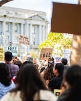 A large group of people gathered in a protest holding various signs advocating for Palestine. The signs display messages such as 'Palestine will be free' and 'No more genocide'. The setting appears to be in front of a large classical-style building with columns.