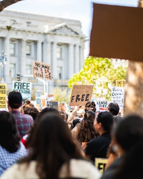 A large group of people gathered in a protest holding various signs advocating for Palestine. The signs display messages such as 'Palestine will be free' and 'No more genocide'. The setting appears to be in front of a large classical-style building with columns.