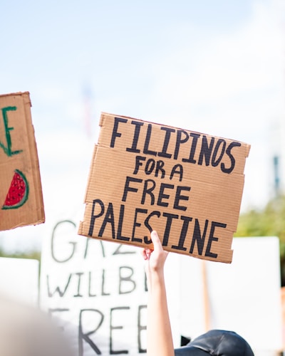 A hand holding up a cardboard sign with the words 'Filipinos for a Free Palestine' written in bold letters. Other protest signs are partially visible in the background, indicating a demonstration. The sky is clear and bright.