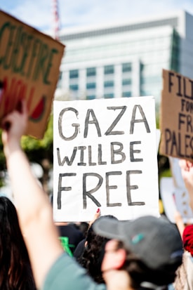 A group of people hold protest signs, prominently featuring a sign with the words 'Gaza Will Be Free' written in bold letters. The scene is set outdoors in what appears to be a public or urban environment with a building in the background.
