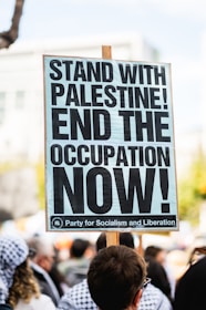 A cardboard sign with bold black text reading 'STAND WITH PALESTINE! END THE OCCUPATION NOW!' is held up by a person in a crowded outdoor setting. There are several people in the background, some wearing patterned headscarves.