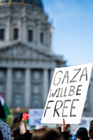 A protest scene with a hand holding a sign reading 'Gaza Will Be Free', set against the backdrop of a large, historic building. The environment suggests an outdoor gathering with other signs and flags visible, indicating a demonstration.