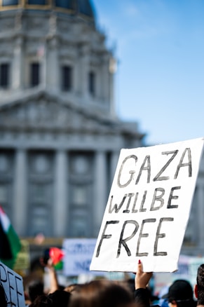 A protest scene with a hand holding a sign reading 'Gaza Will Be Free', set against the backdrop of a large, historic building. The environment suggests an outdoor gathering with other signs and flags visible, indicating a demonstration.