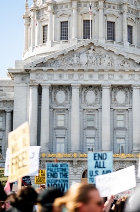 A group of people are holding protest signs in front of a grand, neoclassical building with large columns and detailed architectural features. The focus is on the signs, some of which have messages about ending aid. The American flag is visible on top of the building.