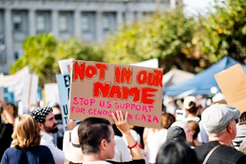 A crowd of people gathered in protest are holding signs. One prominent sign reads 'Not In Our Name, Stop U.S. Support for Genocide in Gaza' with bold red letters. The setting appears to be outdoors in a city area, with trees and a building in the background.