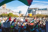 A large crowd gathers in a public square, many holding signs and flags featuring red, green, white, and black colors. The scene is vibrant with people demonstrating in front of a building with flags flying, possibly governmental or civic architecture. Bright sunlight casts shadows across the participants.