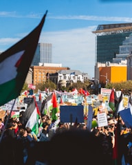A large crowd of protesters gathered in an urban setting, holding numerous signs and flags with messages advocating for peace and justice. The word 'TRUTH' is prominently visible on a building in the background. Various national and political flags are raised amidst the protestors.