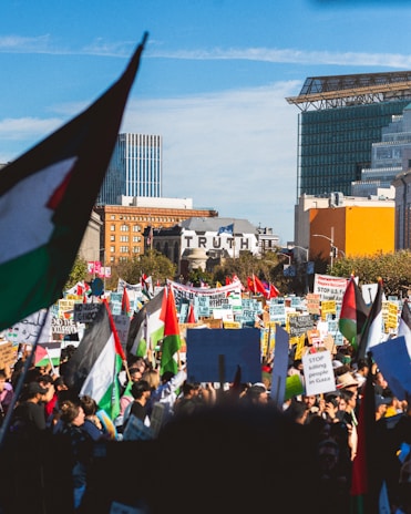 A large crowd of protesters gathered in an urban setting, holding numerous signs and flags with messages advocating for peace and justice. The word 'TRUTH' is prominently visible on a building in the background. Various national and political flags are raised amidst the protestors.