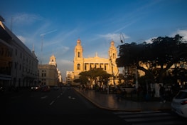 A historic city square is shown at sunset, featuring a prominent yellow cathedral with twin towers and a cross on top. There are people walking and sitting around, with cars driving on the surrounding road. Trees and streetlamps line the pavement, creating shadows against the dusk sky.