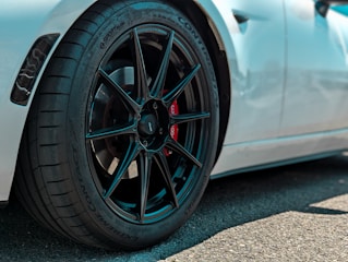Close-up view of a high-performance car wheel featuring a sleek black alloy rim and a red brake caliper. The tire has detailed tread patterns and multiple brand markings. The car's white body is partially visible in the background.