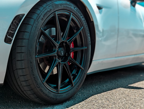 Close-up view of a high-performance car wheel featuring a sleek black alloy rim and a red brake caliper. The tire has detailed tread patterns and multiple brand markings. The car's white body is partially visible in the background.