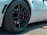 Close-up view of a high-performance car wheel featuring a sleek black alloy rim and a red brake caliper. The tire has detailed tread patterns and multiple brand markings. The car's white body is partially visible in the background.