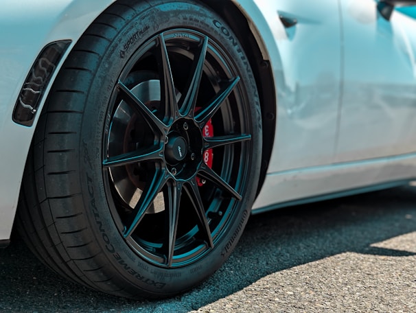 Close-up view of a high-performance car wheel featuring a sleek black alloy rim and a red brake caliper. The tire has detailed tread patterns and multiple brand markings. The car's white body is partially visible in the background.