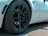 Close-up view of a high-performance car wheel featuring a sleek black alloy rim and a red brake caliper. The tire has detailed tread patterns and multiple brand markings. The car's white body is partially visible in the background.