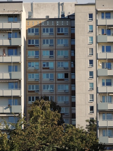 A residential building facade with multiple stories, windowed apartments, and balconies. Trees with dense green foliage are in the foreground, partially obscuring the lower levels of the building.