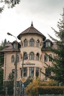 A large, elegant building with a tower-like structure and multiple arched windows is set against a backdrop of trees. The architecture features a light beige facade with brown roof tiles. There is a sign with three stars and some text near the entrance. The surrounding area includes light poles and road signs, adding to the urban setting.
