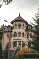 A large, elegant building with a tower-like structure and multiple arched windows is set against a backdrop of trees. The architecture features a light beige facade with brown roof tiles. There is a sign with three stars and some text near the entrance. The surrounding area includes light poles and road signs, adding to the urban setting.