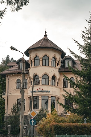 A large, elegant building with a tower-like structure and multiple arched windows is set against a backdrop of trees. The architecture features a light beige facade with brown roof tiles. There is a sign with three stars and some text near the entrance. The surrounding area includes light poles and road signs, adding to the urban setting.