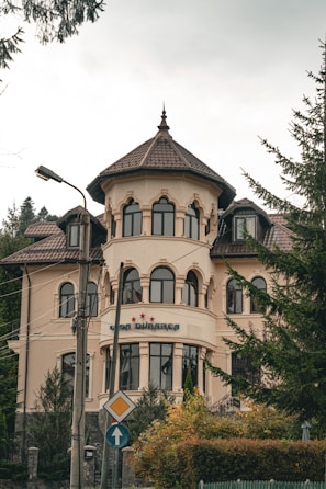 A large, elegant building with a tower-like structure and multiple arched windows is set against a backdrop of trees. The architecture features a light beige facade with brown roof tiles. There is a sign with three stars and some text near the entrance. The surrounding area includes light poles and road signs, adding to the urban setting.