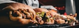 a close-up of several sandwiches lined up on a marble countertop. Each sandwich is filled with various ingredients such as meats, vegetables, and sauces. In the background, there are indistinct kitchen elements, and the image appears to be taken in a restaurant or deli setting.