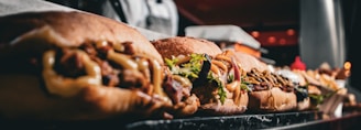 a close-up of several sandwiches lined up on a marble countertop. Each sandwich is filled with various ingredients such as meats, vegetables, and sauces. In the background, there are indistinct kitchen elements, and the image appears to be taken in a restaurant or deli setting.