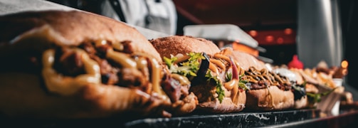 a close-up of several sandwiches lined up on a marble countertop. Each sandwich is filled with various ingredients such as meats, vegetables, and sauces. In the background, there are indistinct kitchen elements, and the image appears to be taken in a restaurant or deli setting.