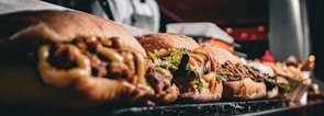 a close-up of several sandwiches lined up on a marble countertop. Each sandwich is filled with various ingredients such as meats, vegetables, and sauces. In the background, there are indistinct kitchen elements, and the image appears to be taken in a restaurant or deli setting.