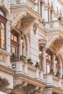 A detailed view of a building facade featuring intricate architectural details, including ornate carvings and decorative columns. The structure showcases classic design elements such as elegant balconies adorned with potted plants, delivering charm and sophistication. Warm sunlight enhances the color of the stonework and wooden window frames.