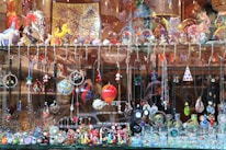 A display window filled with a vibrant array of handcrafted jewelry items and decorative glass figurines. The display includes various necklaces and pendants hanging from hooks, and intricate earrings showcasing different designs and colors. The shelf above features an assortment of colorful glass sculptures, including animals and abstract shapes. The background consists of reflective surfaces and additional decorative items placed in the window.
