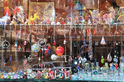 A display window filled with a vibrant array of handcrafted jewelry items and decorative glass figurines. The display includes various necklaces and pendants hanging from hooks, and intricate earrings showcasing different designs and colors. The shelf above features an assortment of colorful glass sculptures, including animals and abstract shapes. The background consists of reflective surfaces and additional decorative items placed in the window.