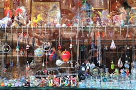 A display window filled with a vibrant array of handcrafted jewelry items and decorative glass figurines. The display includes various necklaces and pendants hanging from hooks, and intricate earrings showcasing different designs and colors. The shelf above features an assortment of colorful glass sculptures, including animals and abstract shapes. The background consists of reflective surfaces and additional decorative items placed in the window.