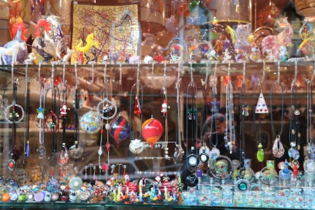 A display window filled with a vibrant array of handcrafted jewelry items and decorative glass figurines. The display includes various necklaces and pendants hanging from hooks, and intricate earrings showcasing different designs and colors. The shelf above features an assortment of colorful glass sculptures, including animals and abstract shapes. The background consists of reflective surfaces and additional decorative items placed in the window.