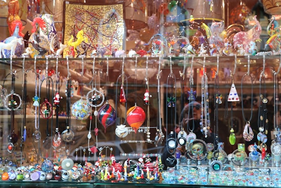 A display window filled with a vibrant array of handcrafted jewelry items and decorative glass figurines. The display includes various necklaces and pendants hanging from hooks, and intricate earrings showcasing different designs and colors. The shelf above features an assortment of colorful glass sculptures, including animals and abstract shapes. The background consists of reflective surfaces and additional decorative items placed in the window.