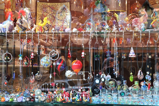 A display window filled with a vibrant array of handcrafted jewelry items and decorative glass figurines. The display includes various necklaces and pendants hanging from hooks, and intricate earrings showcasing different designs and colors. The shelf above features an assortment of colorful glass sculptures, including animals and abstract shapes. The background consists of reflective surfaces and additional decorative items placed in the window.