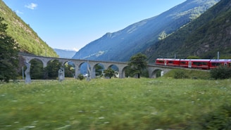 A scenic landscape featuring a red train traveling across a stone viaduct. The viaduct has multiple arches and is flanked by lush green hillsides and a field speckled with grass and some abstract sculptures. The sky above is mostly clear with a few clouds, and the distant mountains complete the picturesque setting.