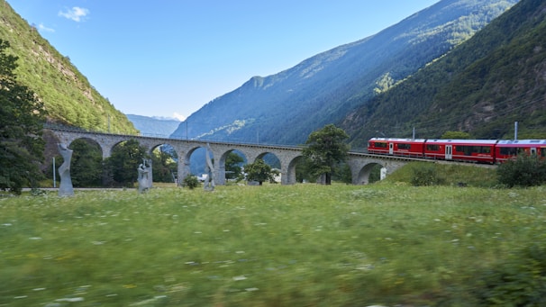 A scenic landscape featuring a red train traveling across a stone viaduct. The viaduct has multiple arches and is flanked by lush green hillsides and a field speckled with grass and some abstract sculptures. The sky above is mostly clear with a few clouds, and the distant mountains complete the picturesque setting.