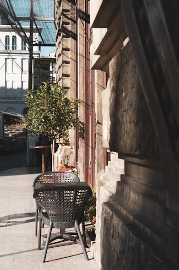An outdoor caf&eacute; setting with two woven chairs and a table, set along a narrow stone-paved pathway adjacent to a building. A small plant and a display of flowers add greenery to the scene. Shadows from overhead structures and the building walls create patterns on the pavement.