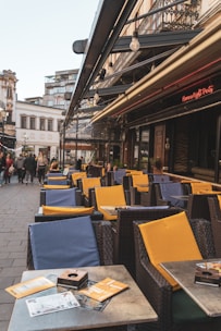 An outdoor café setting with wicker chairs and yellow and blue cushions. Empty tables are lined up along the sidewalk, each with menus or leaflets. Patrons are seen walking in the background on a bustling street with old buildings and ironwork balconies. A warm, inviting atmosphere is complemented by a neon sign on the awning.