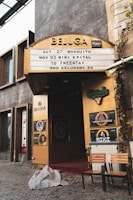 An entrance to a venue named Beluga with a signage displaying upcoming events and dates. The entrance is framed with golden panels and has various posters including Carlsberg and Trooper Friends. Two chairs and a small table are placed outside on a cobblestone pavement with a blanket lying on the ground.