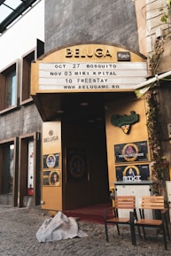 An entrance to a venue named Beluga with a signage displaying upcoming events and dates. The entrance is framed with golden panels and has various posters including Carlsberg and Trooper Friends. Two chairs and a small table are placed outside on a cobblestone pavement with a blanket lying on the ground.