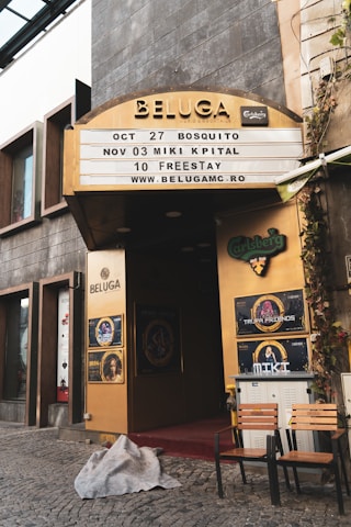 An entrance to a venue named Beluga with a signage displaying upcoming events and dates. The entrance is framed with golden panels and has various posters including Carlsberg and Trooper Friends. Two chairs and a small table are placed outside on a cobblestone pavement with a blanket lying on the ground.