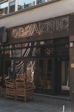 A dimly lit cafe exterior with the name 'Obraznic' displayed in neon lights above the entrance. The door and windows are dark, with reflections visible on the glass. Several wooden chairs are stacked outside on the pavement, suggesting they may be used for outdoor seating.