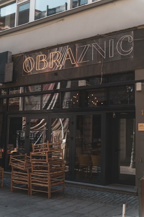 A dimly lit cafe exterior with the name 'Obraznic' displayed in neon lights above the entrance. The door and windows are dark, with reflections visible on the glass. Several wooden chairs are stacked outside on the pavement, suggesting they may be used for outdoor seating.