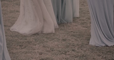 Flowing dresses made of light, delicate fabric gently cascade to the ground, covering the grass. The colors of the dresses range from muted blues to soft greens, creating a serene and ethereal atmosphere.