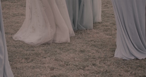 Flowing dresses made of light, delicate fabric gently cascade to the ground, covering the grass. The colors of the dresses range from muted blues to soft greens, creating a serene and ethereal atmosphere.