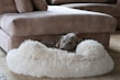 A dog is comfortably sleeping in a fluffy beige dog bed positioned next to a brown suede sofa. The setting appears cozy and calm, with soft furnishings and muted tones creating a peaceful atmosphere.
