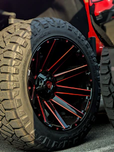 Close-up of a large off-road tire with aggressive tread patterns, mounted on a black and red spoked rim. The wheel is part of a vehicle with a visible portion in bright red.