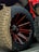 Close-up of a large off-road tire with aggressive tread patterns, mounted on a black and red spoked rim. The wheel is part of a vehicle with a visible portion in bright red.