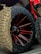 Close-up of a large off-road tire with aggressive tread patterns, mounted on a black and red spoked rim. The wheel is part of a vehicle with a visible portion in bright red.